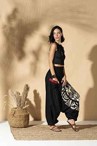 Woman wearing black outfit with patterned pants standing on a rug next to a basket with tall grass.